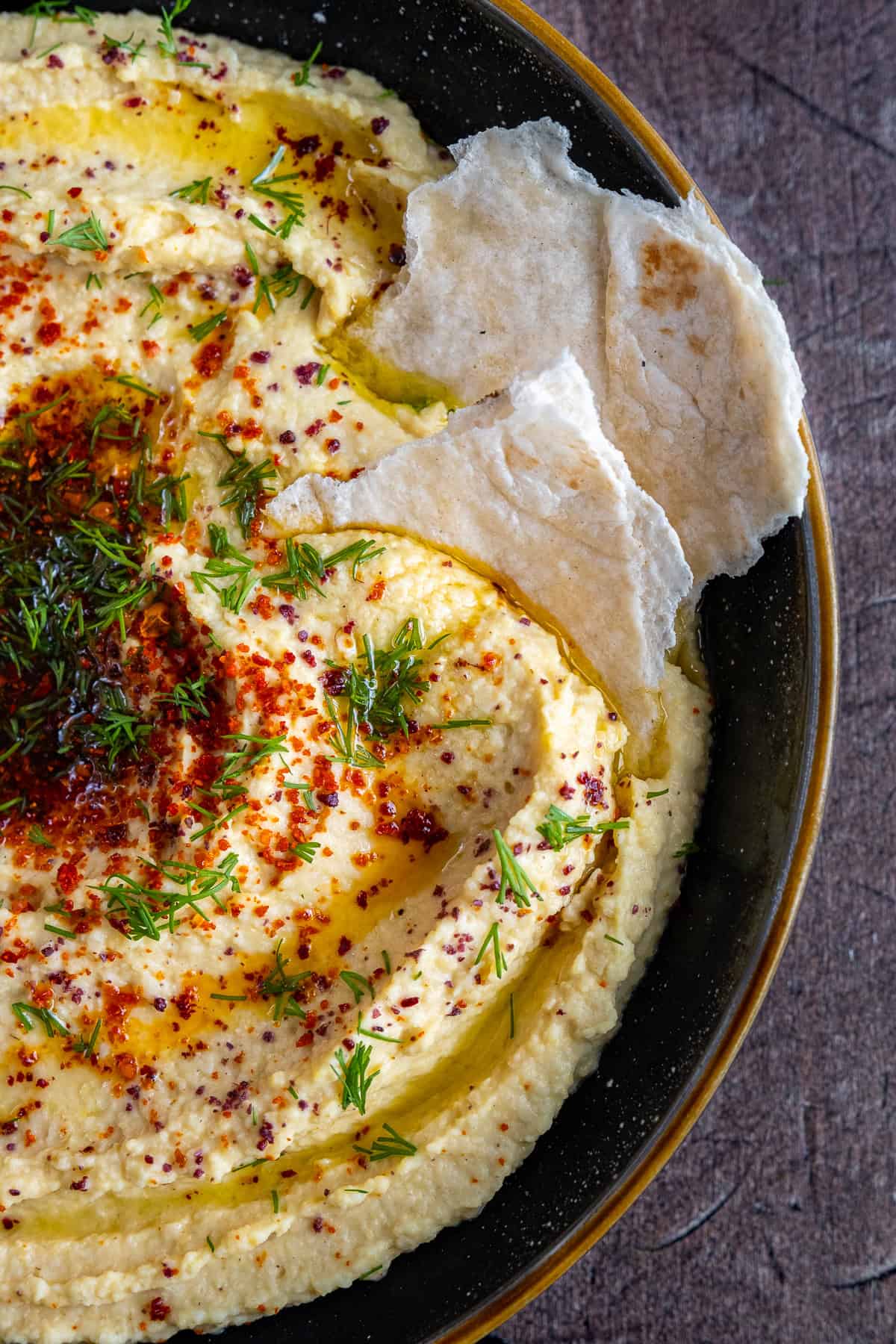 A close-up shot of hummus garnished with red pepper flakes, sumac, and fresh dill, with two pieces of torn flatbread dipped into it. 