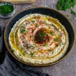 Hummus garnished with red pepper flakes, sumac and fresh dill served in a black bowl.
