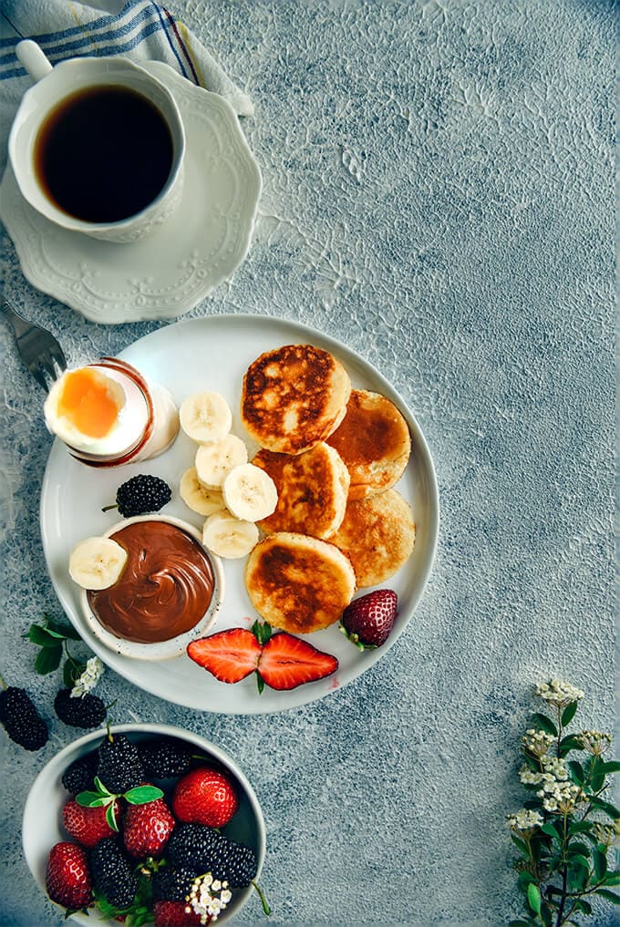 Yogurt pancakes with berries, banana slices, chocolate and a soft boiled egg on a white plate accompanied by a cup of tea for breakfast