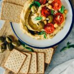 Hummus without tahini garnished with olives, tomatoes, herbs and sun flower seeds served with crackers.