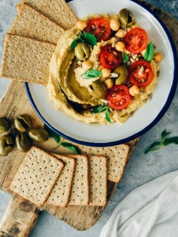 Hummus without tahini garnished with olives, tomatoes, herbs and sun flower seeds served with crackers.