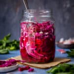 A jar of pickled red cabbage on a dark background, herbs and garlic cloves on the side.