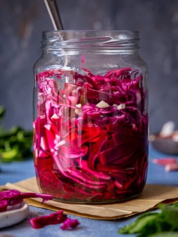 A jar of pickled red cabbage on a dark background, herbs and garlic cloves on the side.