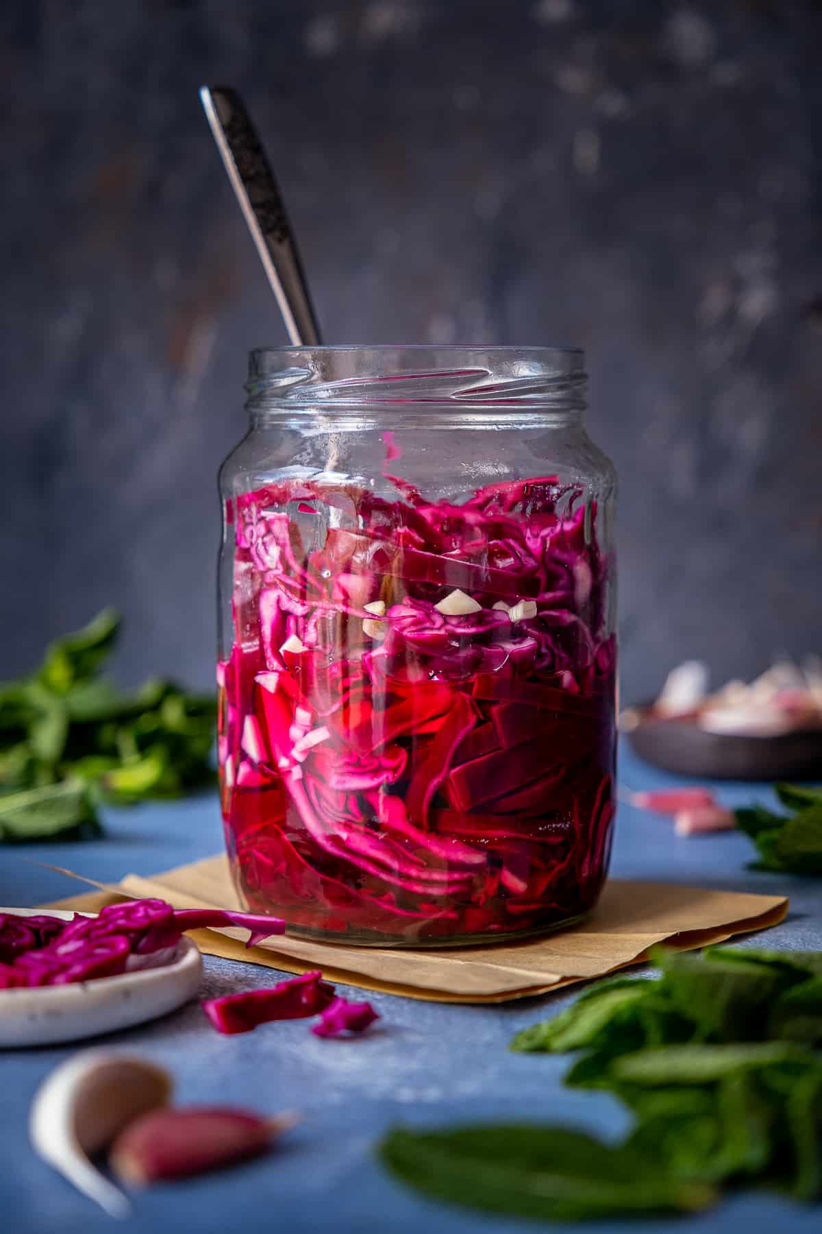 A jar of pickled red cabbage, herbs and garlic cloves on the side on a dark background.