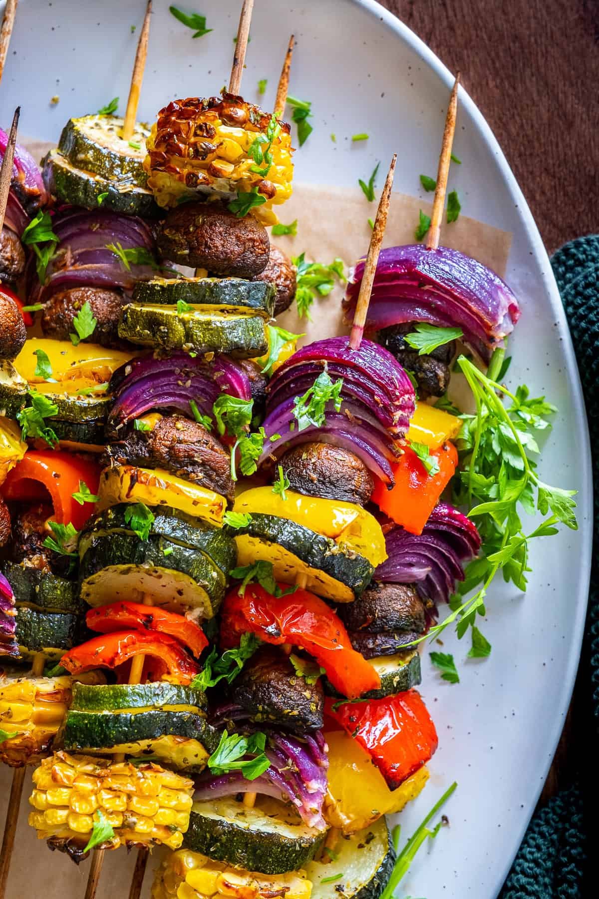 Colorful vegetable kabobs in oven served on a white plate, featuring grilled zucchini, red onion, corn, mushrooms, and bell peppers, garnished with fresh parsley.