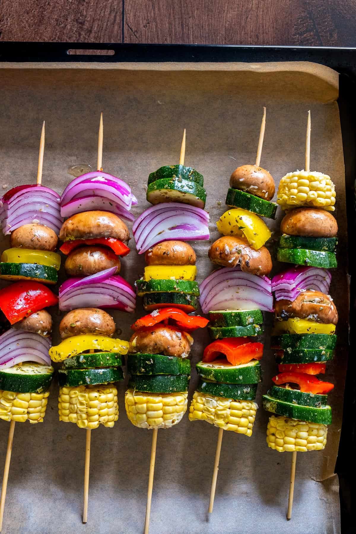 Uncooked vegetable kabobs on wooden skewers arranged on a parchment-lined baking sheet, ready to be roasted in the oven.