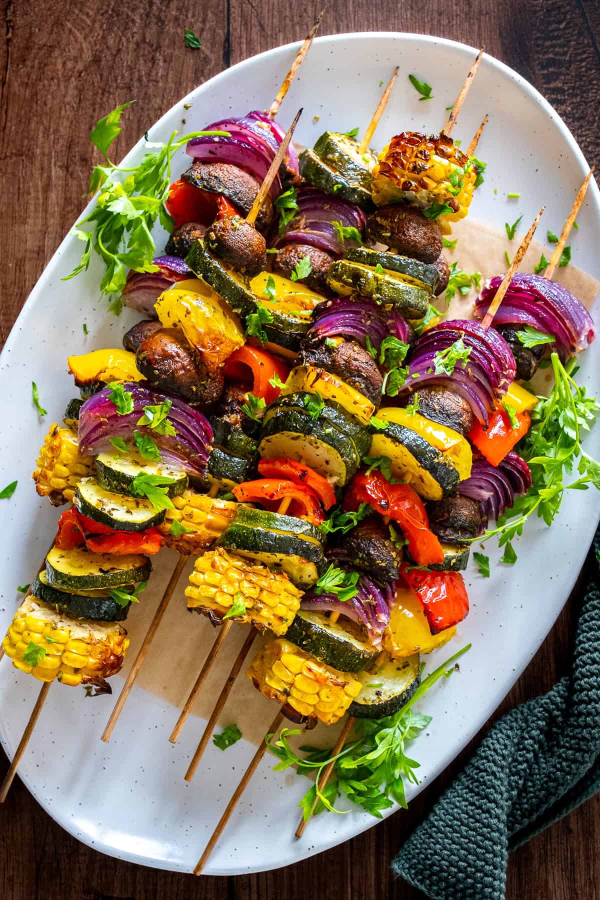 Oven-roasted vegetable kabobs on skewers with zucchini, corn, red onion, bell peppers, and mushrooms, served on a white platter with fresh herbs.