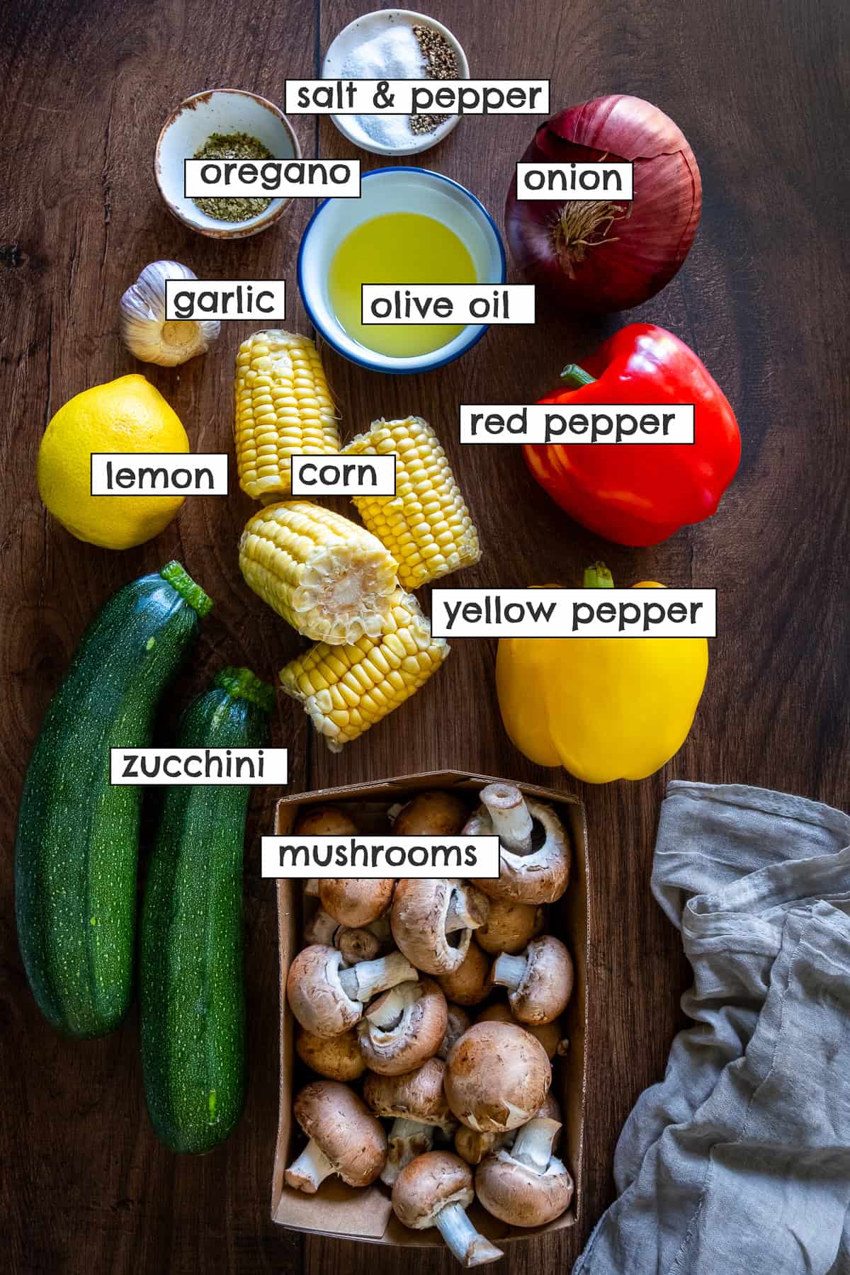 Labeled flat lay of ingredients for vegetable kabobs in oven, including zucchini, mushrooms, corn, bell peppers, onion, garlic, lemon, olive oil, oregano, salt, and pepper.