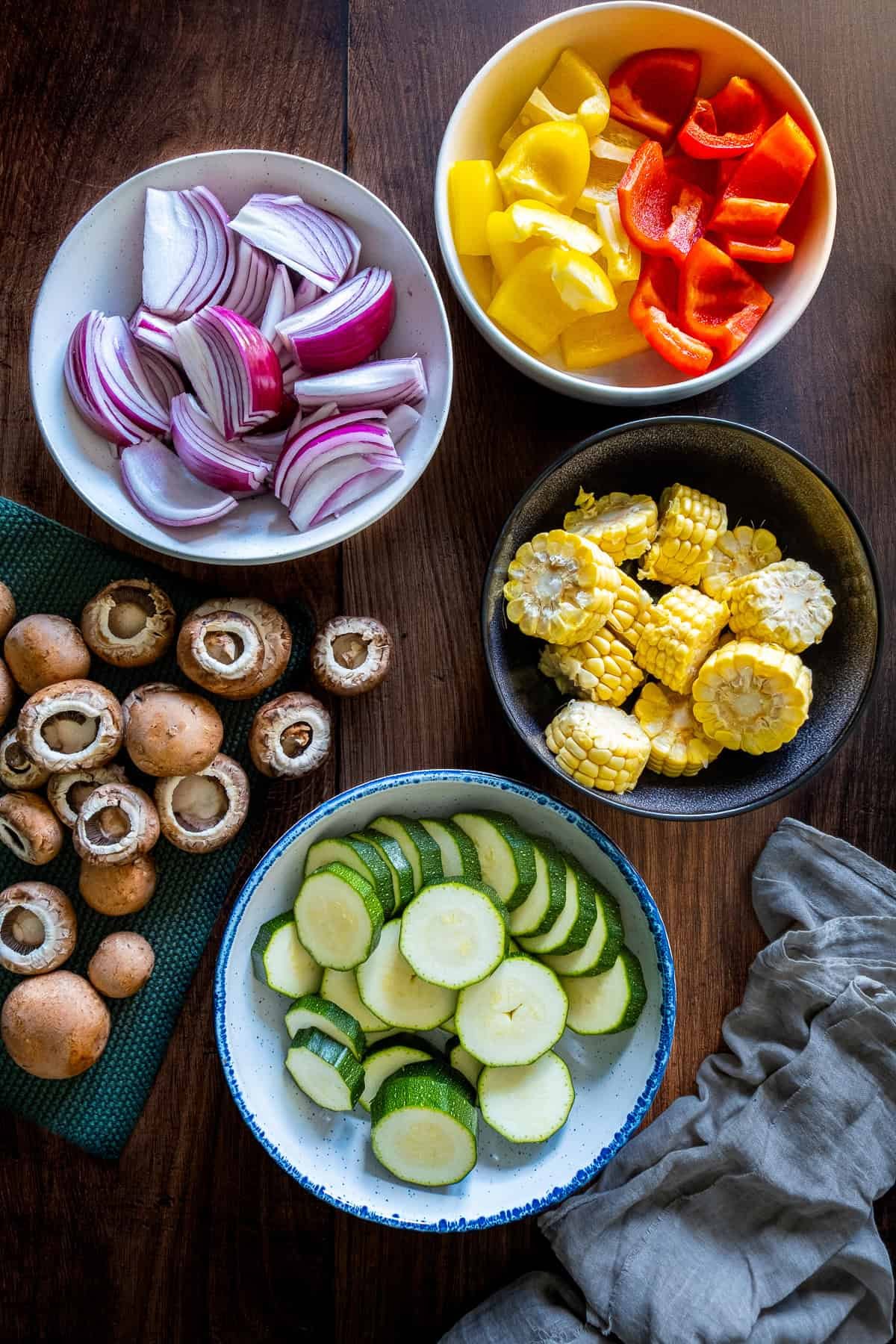 Chopped vegetables for kabobs, including zucchini, red and yellow bell peppers, red onion, corn rounds, and whole mushrooms, arranged in bowls on a wooden surface.