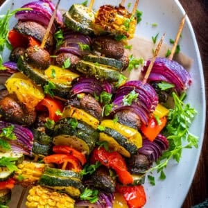 Oven-roasted vegetable kabobs with zucchini, mushrooms, bell peppers, red onions, and corn, garnished with parsley on a white plate.