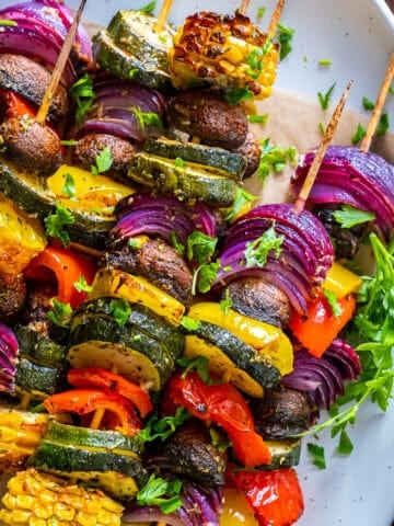 Oven-roasted vegetable kabobs with zucchini, mushrooms, bell peppers, red onions, and corn, garnished with parsley on a white plate.
