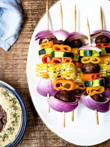 Colorful veggie kabobs on a white plate accompanied by a blue napkin and hummus on the side.