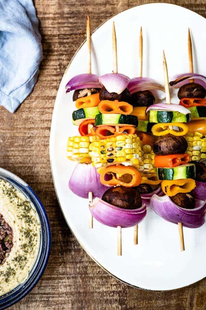 Colorful veggie kabobs on a white plate accompanied by a blue napkin and hummus on the side.