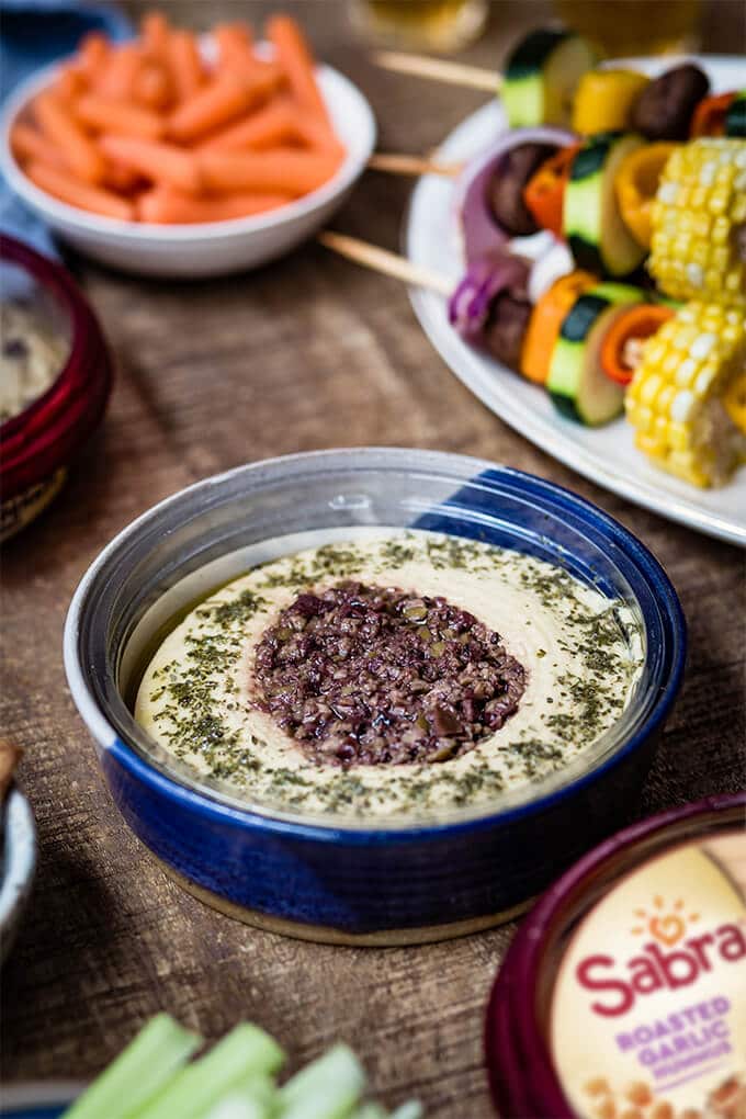 Veggie Kabobs with a simple tangy marinade are so tasty and healthy. So vibrant and packed with flavors, these kabobs make a a great side dish to any meal or a perfect summer appetizer. #vegetarian #kabobs #summerparty #partyfood #meatless #appetizer #ad @sabradips Hummus with olive tapenade in a blue bowl accompanied by veggie shish kabob plate.
