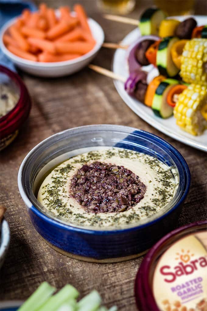 Hummus with olive tapenade in a blue bowl accompanied by veggie shish kabob plate.