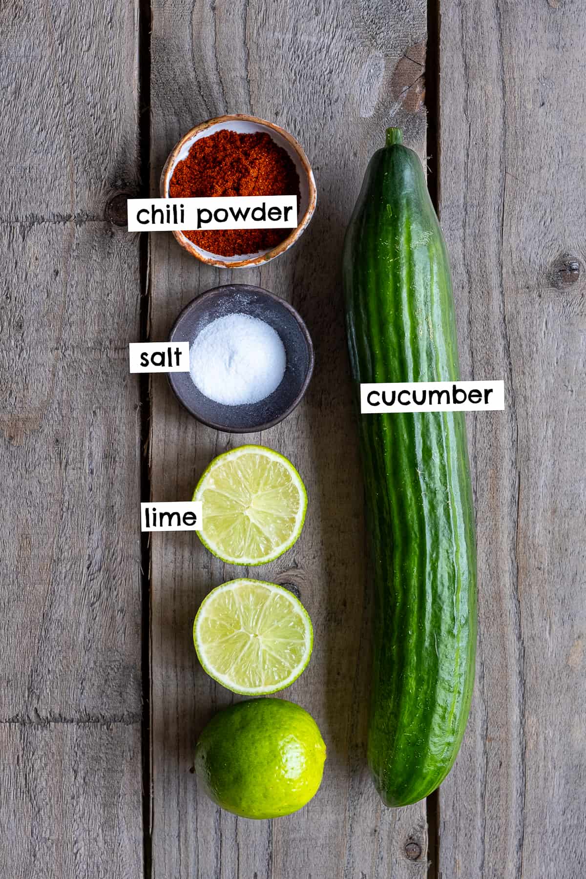 A large cucumber, chili powder, salt, limes on a wooden background.