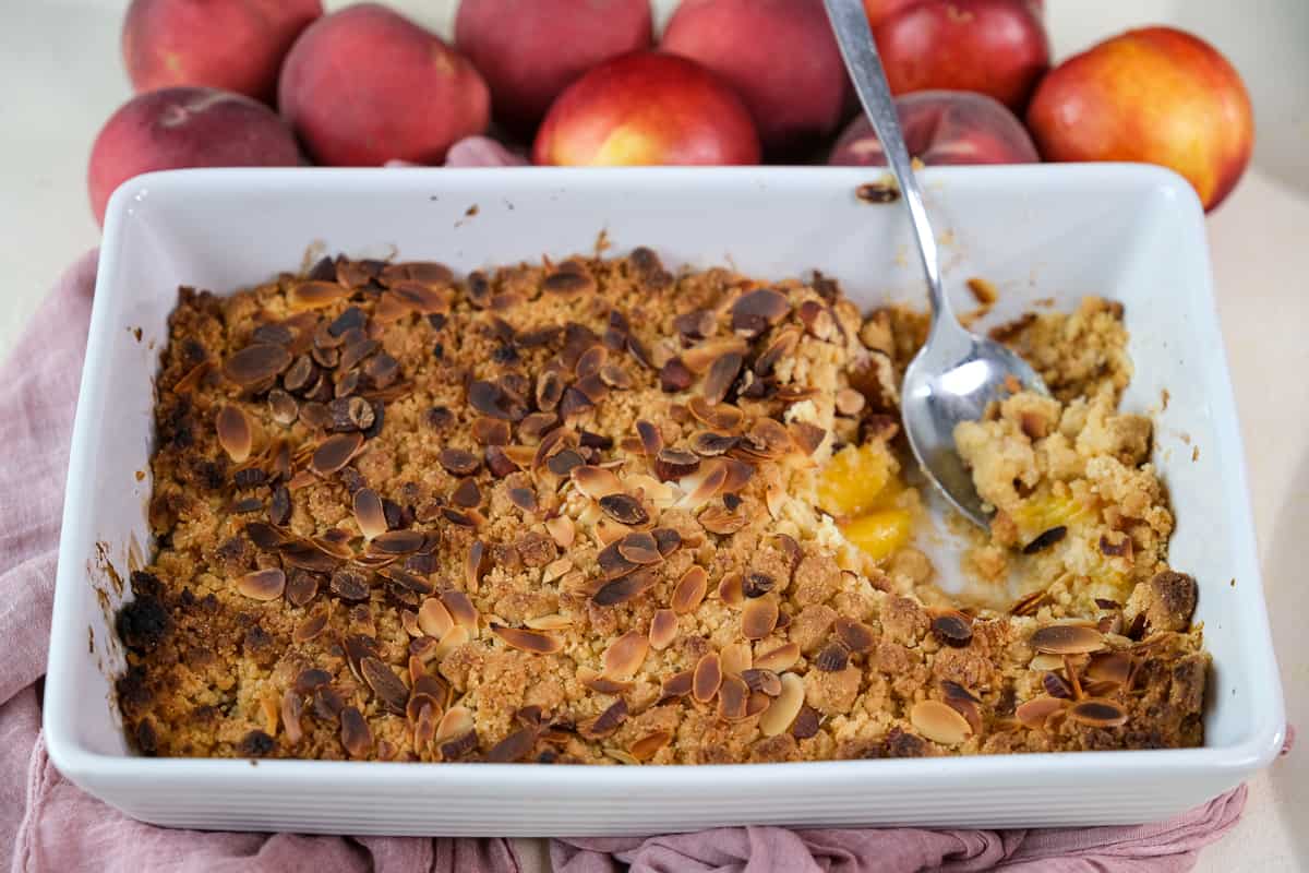 Peach cobbler with cake mix topped with almonds in a white baking pan with a spoon in it and fresh peaches behind it.