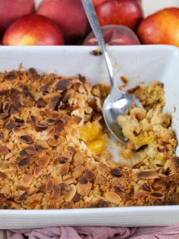 Peach cobbler with cake mix topped with almonds in a white baking pan with a spoon in it and fresh peaches behind it.