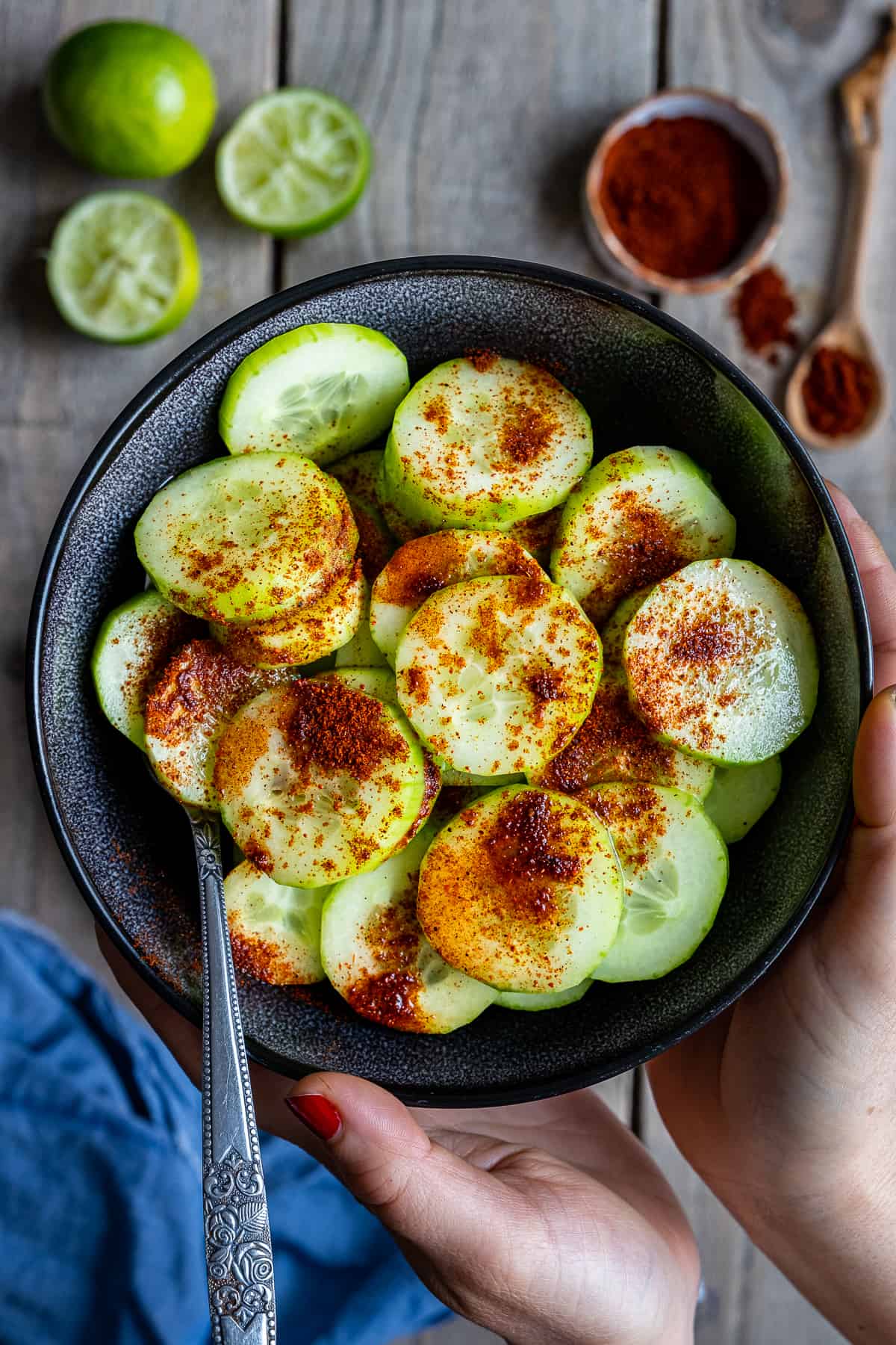 Hands holding a black bowl of cucumber slices topped with chili powder.