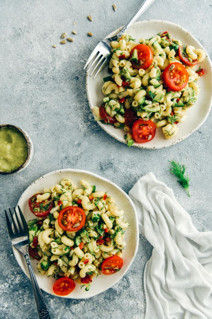 Vegan pasta salad served on two white ceramic plates with a fork on the side of each. Avocado dressing in a small bowl accompany.