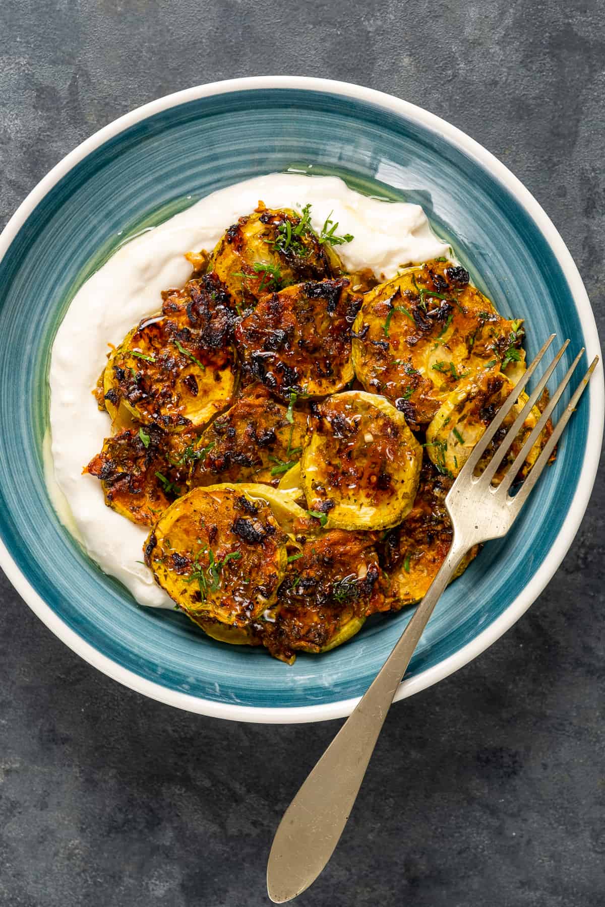 Baked zucchini with tomatoes and herbs served on some yogurt in a blue ceramic dish and a fork inside.