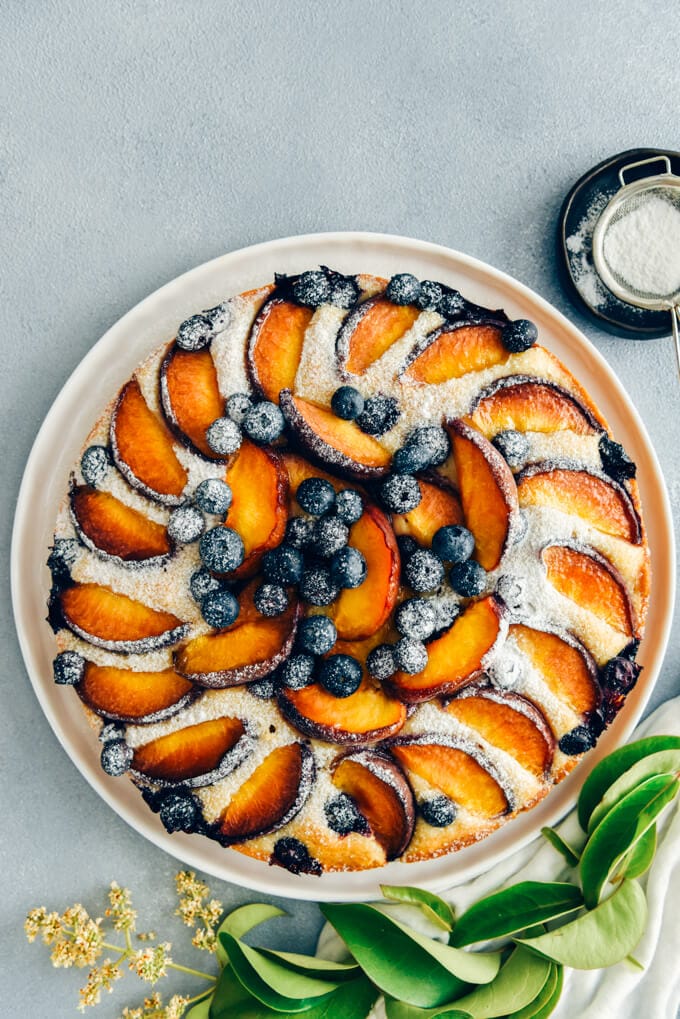 This Blueberry Peach Coffee Cake is a luscious cake that is sure to be a crowd pleaser. Made with olive oil and kefir, this simple cake is moist, tender and so light. #peachcake #peachcoffeecake #peachrecipe #coffeecake #kefircake #kefirrecipes Blueberry peach coffee cake dusted with powdered sugar on a white plate