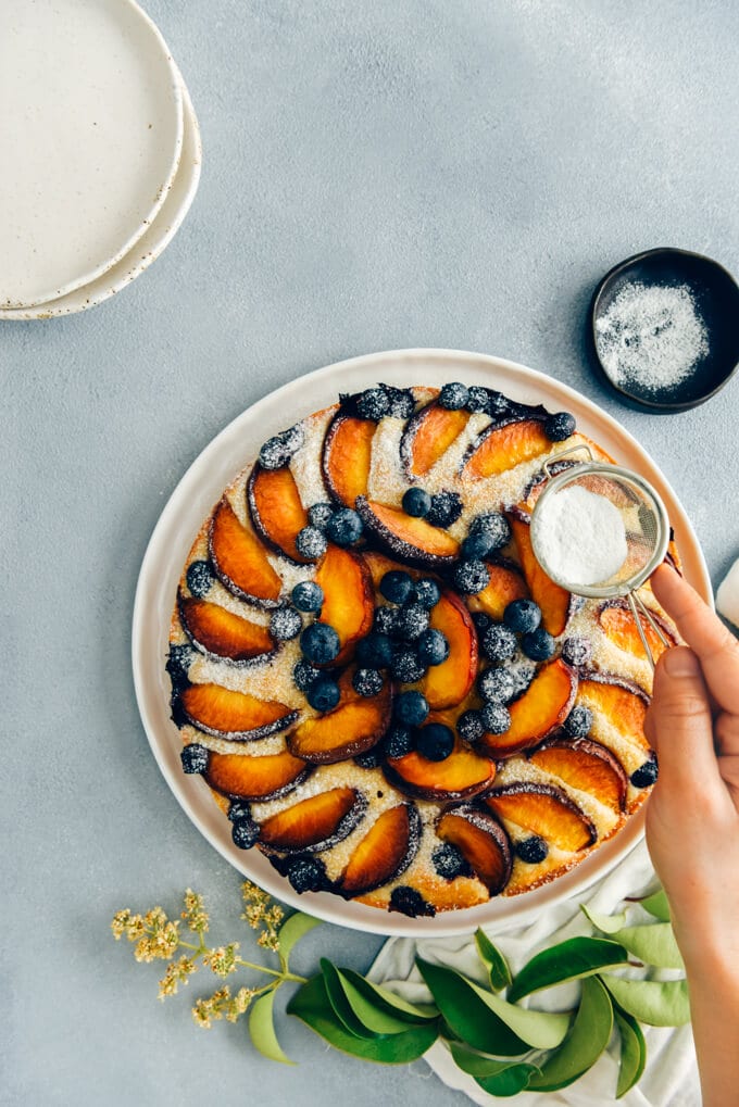 This Blueberry Peach Coffee Cake is a luscious cake that is sure to be a crowd pleaser. Made with olive oil and kefir, this simple cake is moist, tender and so light. #peachcake #peachcoffeecake #peachrecipe #coffeecake #kefircake #kefirrecipes Sifting powdered sugar on peach coffee cake with fresh peaches