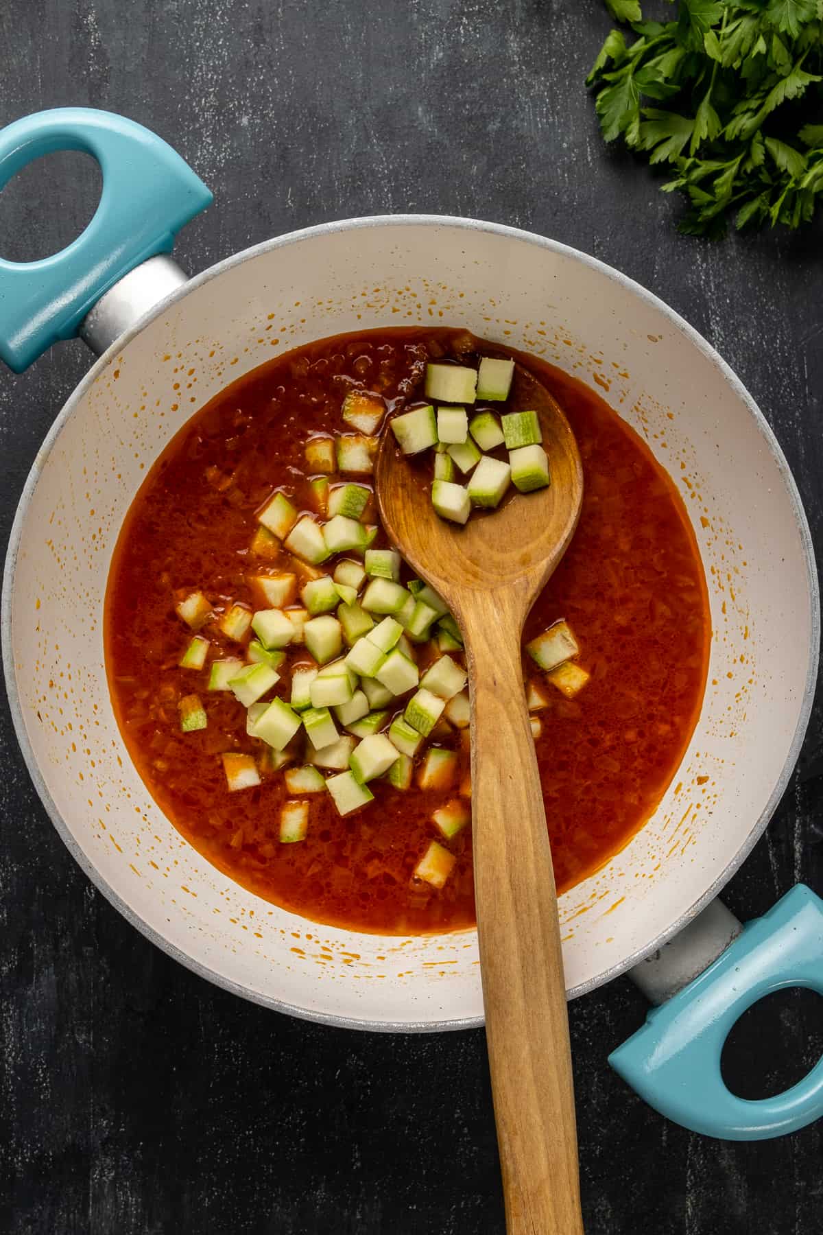 Diced zucchini cooking in a tomato sauce in a pan and a wooden spoon in it.