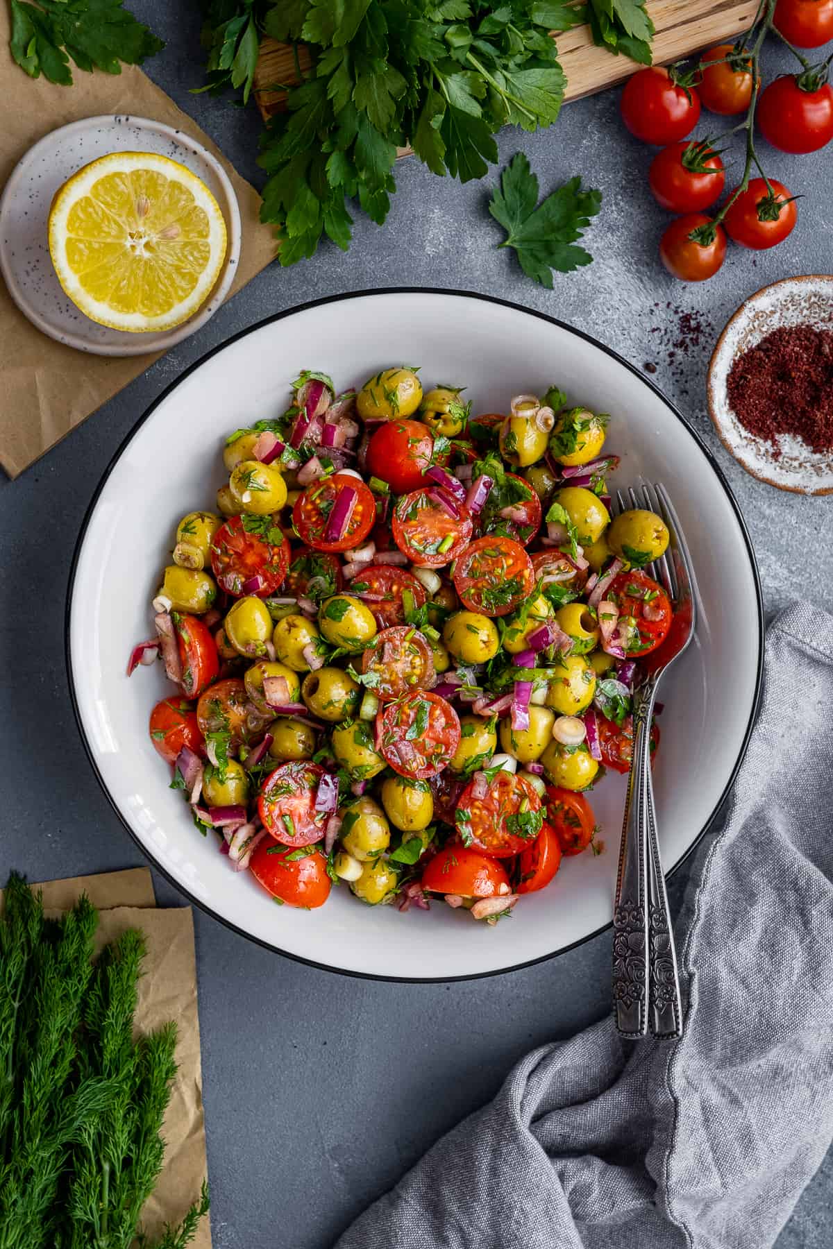 A salad with green olives, cherry tomatoes, onions and herbs in a white bowl and two forks inside it.