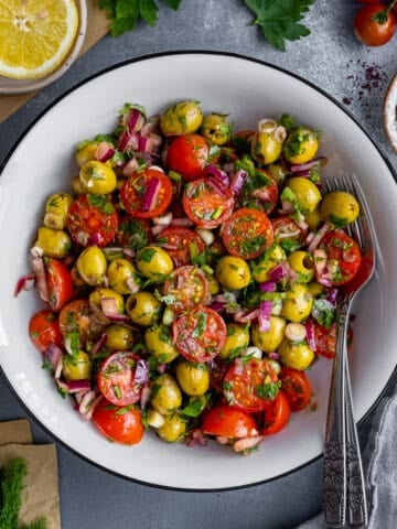 Green olive salad with tomatoes, onions and herbs in a white bowl and two forks inside it.