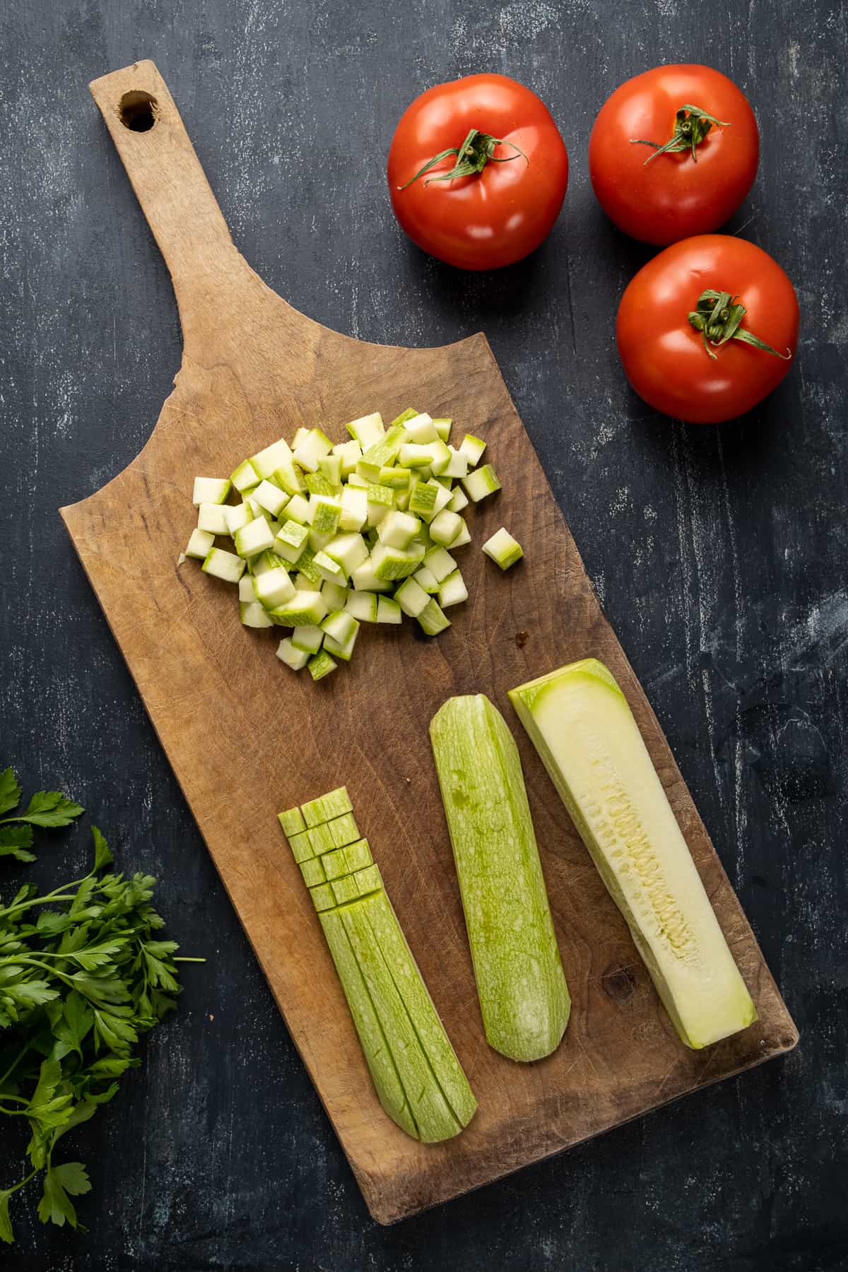 Zucchini sliced and diced on a wooden board, tomatoes and parsley on the side.