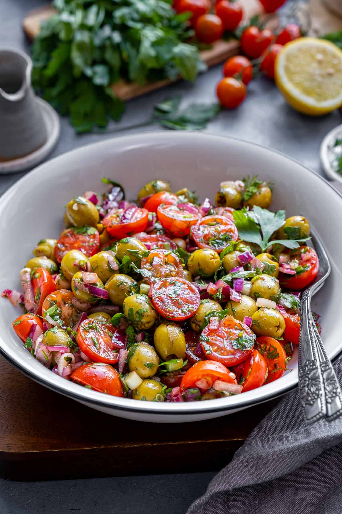 Green olive salad in a white bowl, two forks inside it and lemon, cherry tomatoes and parsley behind it.