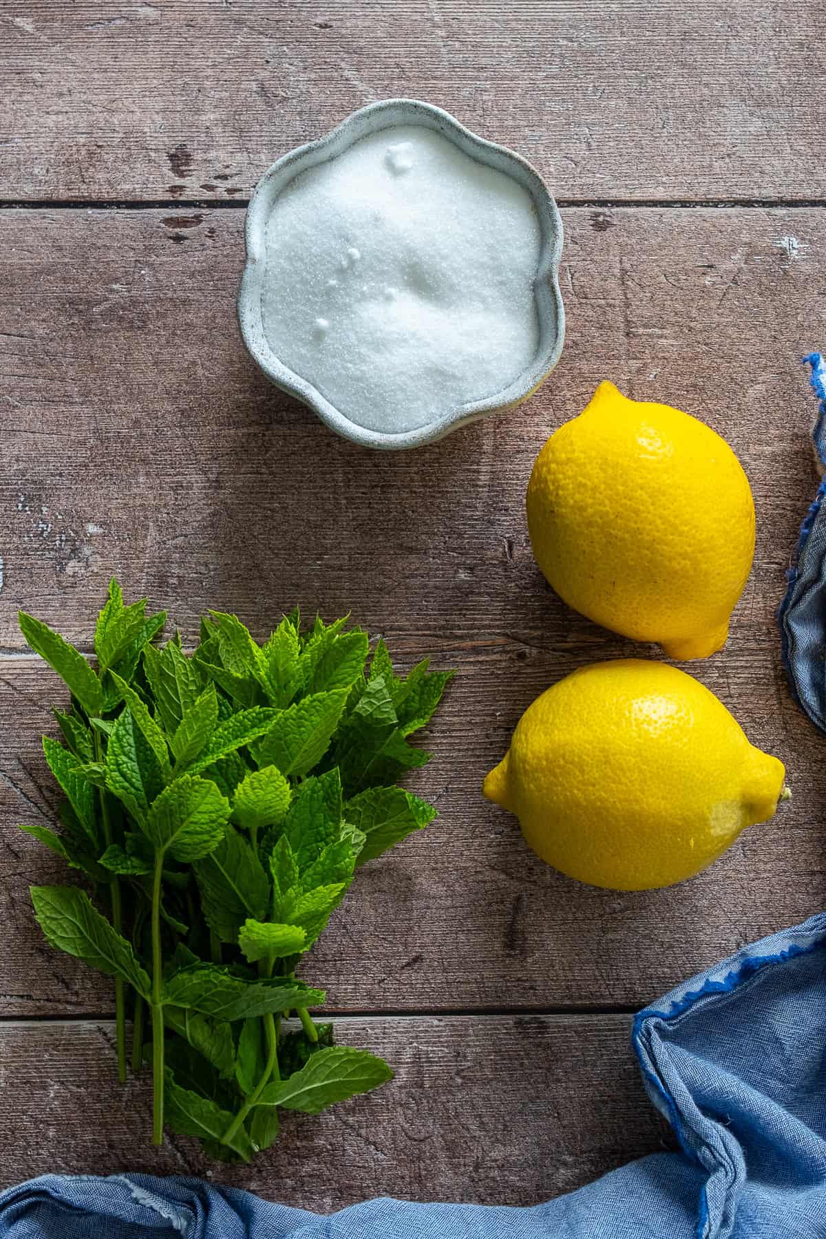 Fresh mint leaves, lemons, and a bowl of sugar on a wooden surface, ingredients for homemade mint lemonade.