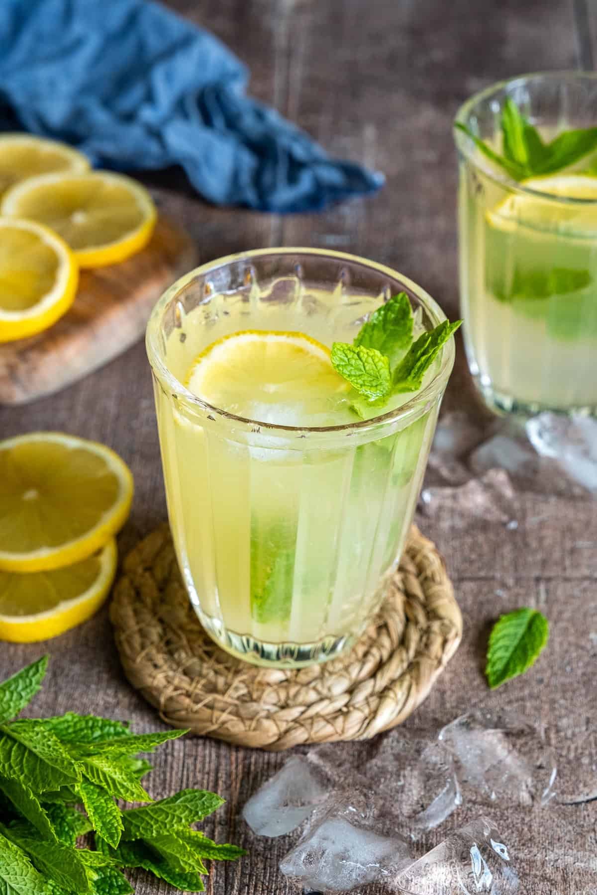A glass of mint lemonade with ice, lemon slices, and fresh mint leaves, served on a rustic table with extra lemon and mint on the side.