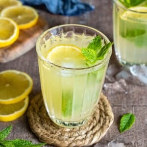 A glass of mint lemonade with lemon slice, mint leaves, and ice cubes on a woven coaster.