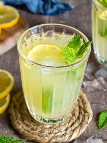 A glass of mint lemonade with lemon slice, mint leaves, and ice cubes on a woven coaster.