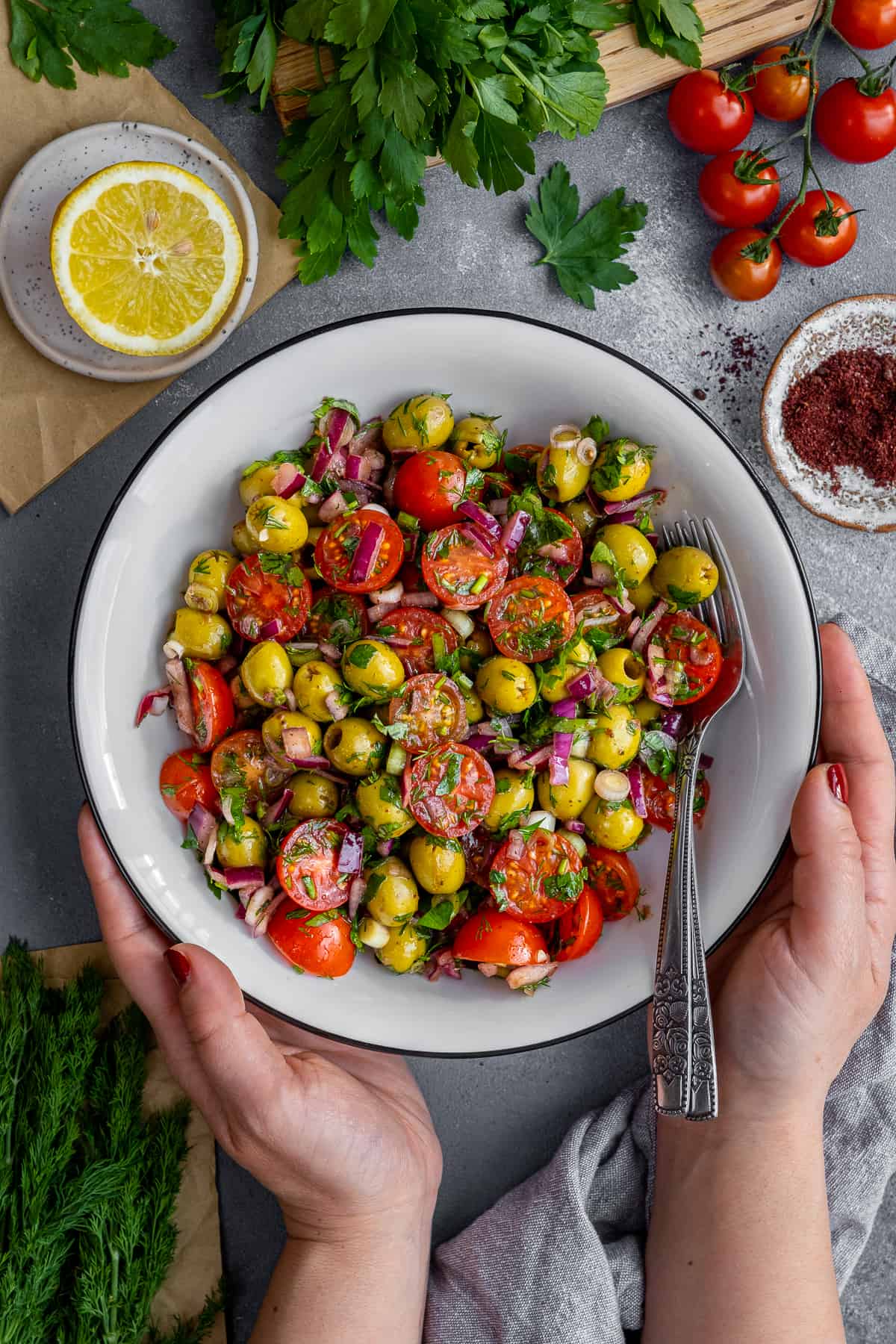 Hands holding green olive salad with tomatoes in a white bowl.