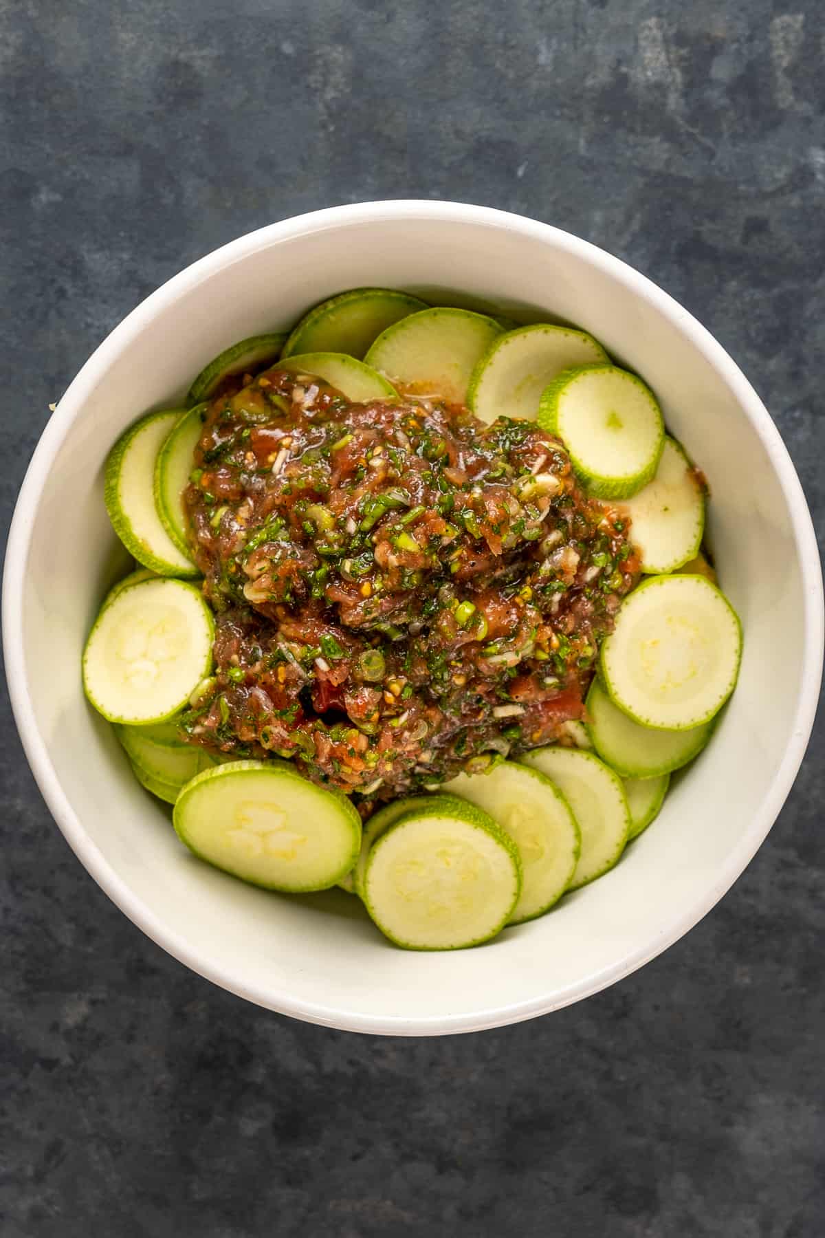 Thin zucchini slices in a white bowl and the tomato puree sauce with herbs poured over them.