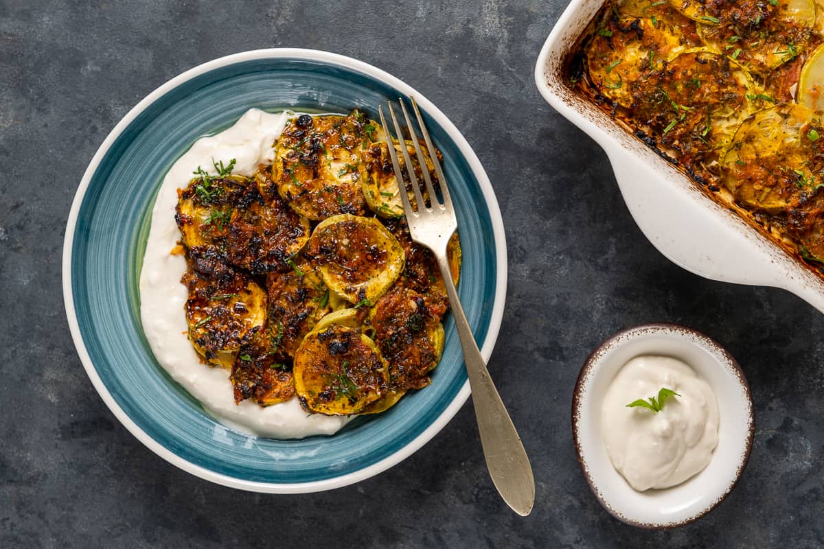 Zucchini casserole served on some yogurt in a blue dish, a fork in it, more yogurt in a small bowl and the rest of the casserole in the baking pan on the side.