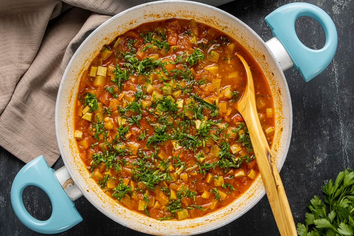 Zucchini tomato stew topped with chopped parsley in a pan and a wooden spoon in it.