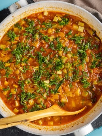 Zucchini stew with tomatoes and herbs in a white pan and a wooden spoon in it.