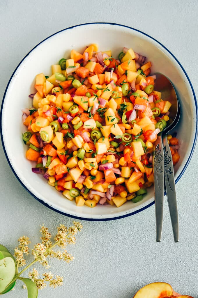 Fresh Corn Peach Salsa is a great recipe to bring summer on your table. Thanks to tangy garden tomatoes, hot peppers and red onion, this is not an overly-sweet salsa. Perfect on chips, chicken, fish or even in sandwiches. #salsa #peachsalsa #salsarecipe #freshpeach #partyfood Corn peach salsa in a white bowl with spoons