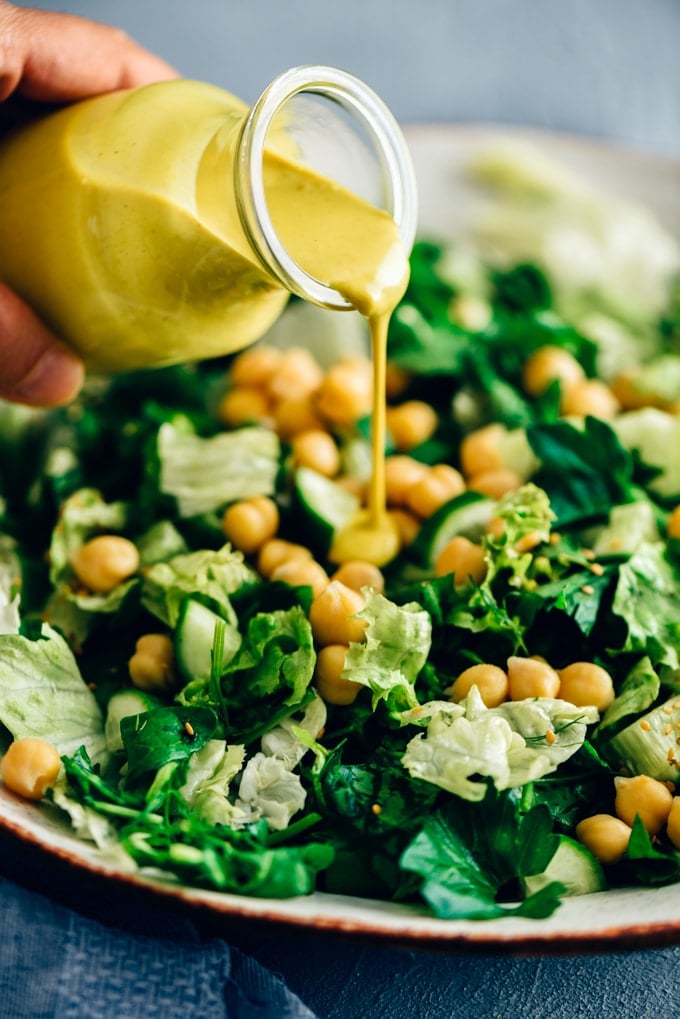 A woman pouring turmeric tahini dressing over a bowl of green salad with chickpeas.