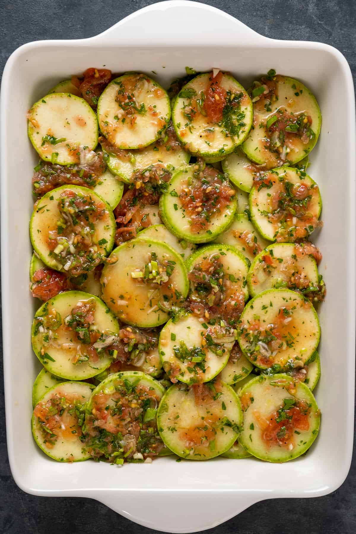Zucchini slices coated with the tomato and herbs sauce in a white rectangular baking pan.