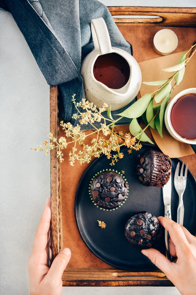 Person holding a wooden tray with healthy chocolate zucchini muffins on a black plate and tea