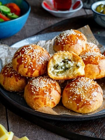 Golden brown pogaca with sesame seeds, one cut open showing a feta and herb filling, served with tea, olives, and cheese.