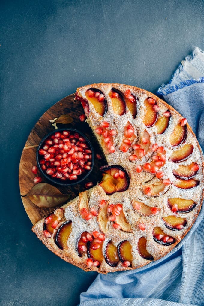 Apple plum cake on a wooden stand accompanied by a small bowl of pomegranate arils.