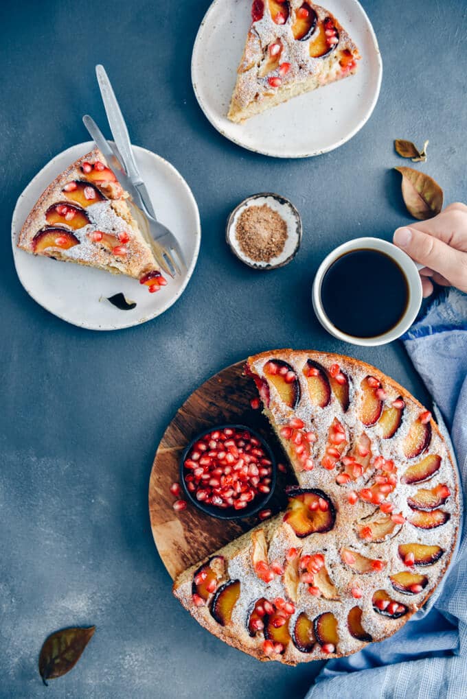 Apple plum cake topped with slices of apple and plum. Cake slices on two white plates, a cup of coffee and a small of brown sugar accompany.