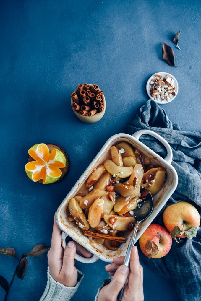 Cinnamon baked apple slices in a baking pan accompanied by apples, cinnamon sticks, orange and almonds.