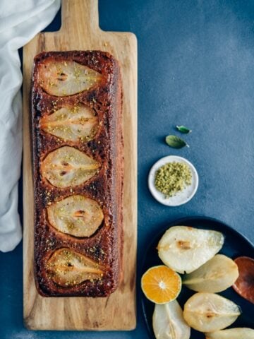 Pear bread on a wooden board accompanied by caramelized pears and a wedge of orange in an iron pan on the side.